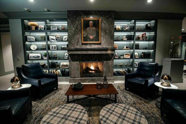The fireplace space in the lobby of Morris Inn. Navy blue leather chairs, plaid ottomans, and a wooden table fill in the space. A fireplace and built-inns with various books and items line the wall.