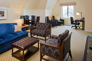 Morris Inn Suite 400 - A blue velvet couch, leather ottoman, and two patterned chairs fill the main space of the room. A brown table with brown chairs fills the back of the room. Two blue armchairs and a desk with an office chair fill a nook near the window