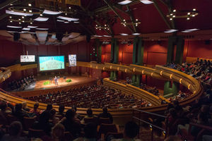 DeBartolo Performing Arts Center - a performer stands on the stage with televisions showing graphics in the background. A large crowd fills all levels of the center with dim, red lighting.