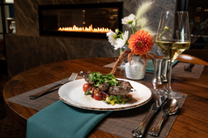 Rohr's Dinner - Meat decorated in greens on top of fresh vegetables sits on a white plate and green tablecloth. A glass of white wine and fresh flowers sit on the table in front of the fireplace.